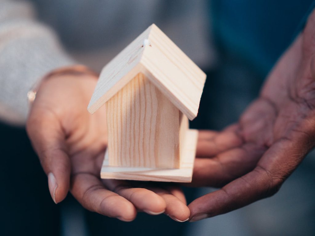 person-holding-miniature-wooden-house-7579128 Close-up of hands holding a wooden house model representing real estate or new home purchase.